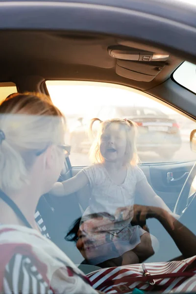 Kleinkind-Mädchen und ihre Mutter im Auto sitzend und lachend, Sonnenuntergang fotografiert mit dem Spiegelbild der Fotografen im Autofenster lizenzfreie Stockbilder