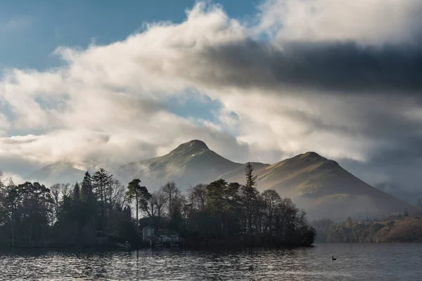 Epic landscape image looking across Derwentwater in Lake District towards Catbells snowcapped mountain with thick fog rolling through valley