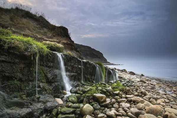 Landscape image of wide waterfall flowing onto rocky beach at su — Stock Photo, Image