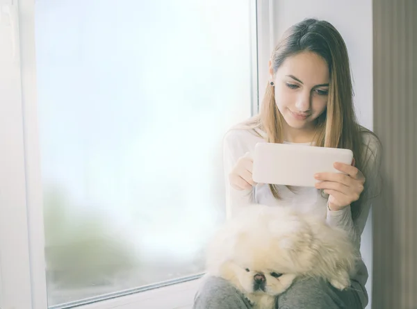 她坐在窗台上和使用 tablet pc 在家里的狗的漂亮女孩. — 图库照片