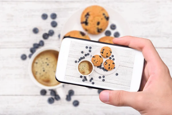 Handen nemen foto blueberry muffins met kop koffie met smartphone. — Stockfoto