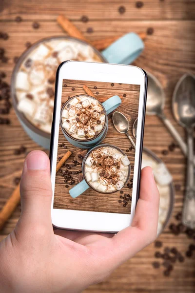 Ruce s foto hrnek kakaa s marshmallows a čokolády s smartphone. — Stock fotografie