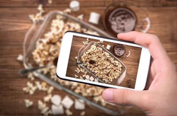 Ruce s foto tmavě čokoládové karamelový popcorn s smartphone. — Stock fotografie