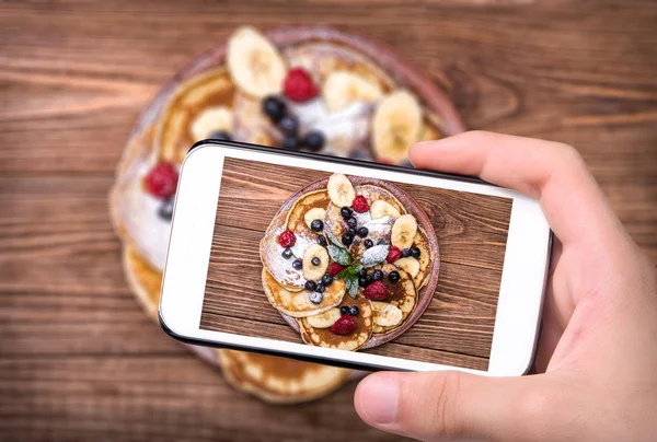 Mãos a tirar fotos de panquecas com fruta fresca com smartphone . — Fotografia de Stock