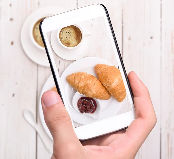 Hands taking photo cup of coffee with croissants with smartphone — Stock Photo, Image