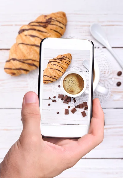 Руки делают фото чашки кофе с круассаном со смартфоном . — стоковое фото
