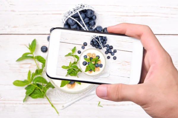 Mãos tirando foto iogurte com muesli e mirtilos com smartphone . — Fotografia de Stock