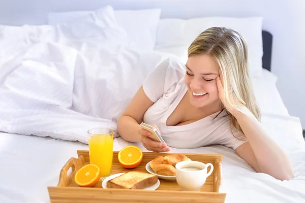 携帯電話を使用しながら、ベッドで朝食を食べる女性. — ストック写真