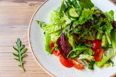 Beyaz tabakta domates, salatalık, soğan, ıspanak, marul ve roka ile taze sebze salatası. Tahta masada sebzeli bir kase salata..