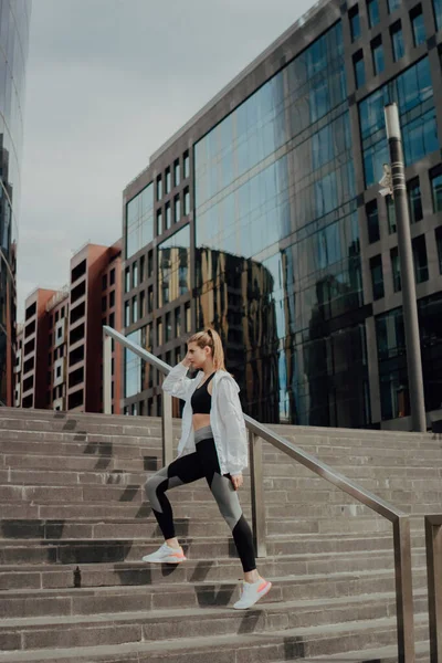 Een jong meisje in een trainingspak staat op de trap tegen de stad — Stockfoto