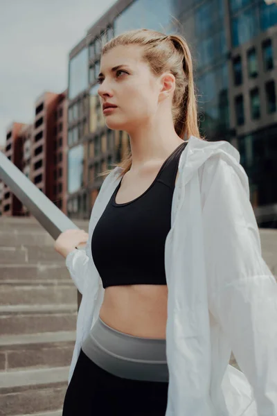 Een jong meisje in een trainingspak staat op de trap tegen de stad — Stockfoto