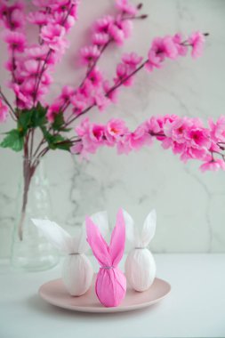 Üç yumurta tavşanı renkli kağıt bir tabakta, kiraz çiçeklerinin yanında. Paskalya tatili masada.