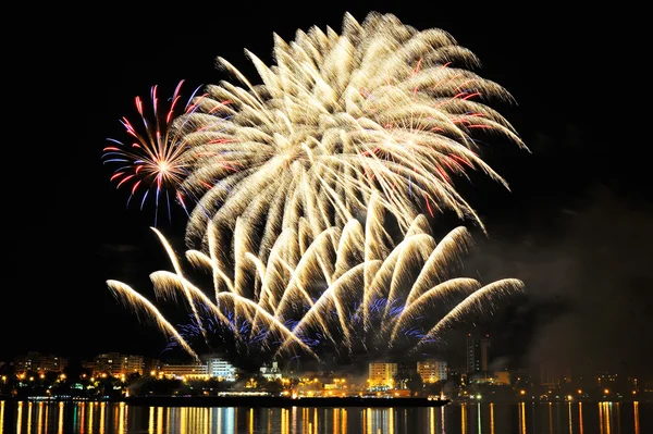 Fogos de artifício sobre a cidade à noite com reflexão na água — Fotografia de Stock