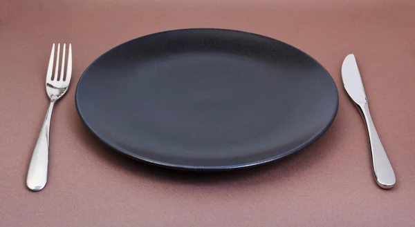 Leerer schwarzer Teller mit Gabel und Messer auf braun — Stockfoto