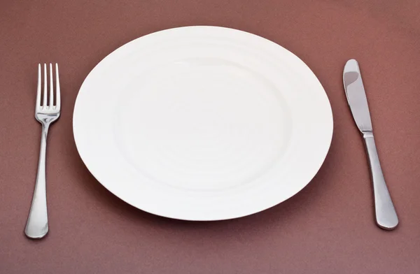 Empty white plate with fork and knife set on brown — Stock Photo, Image