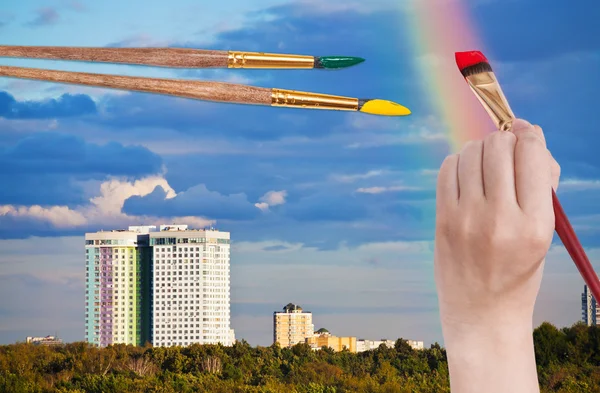 Brush paints rainbow in blue clouds over houses — Stock Photo, Image