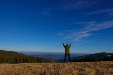 Yürüyüşçü elleri havada, mavi gökyüzü olan geniş bir tepenin üzerinde duruyor.