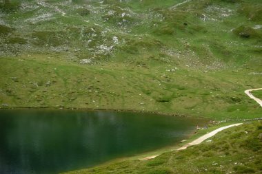 Dağ gölü ve iki yürüyüşçü su manzarasında duruyorlar.