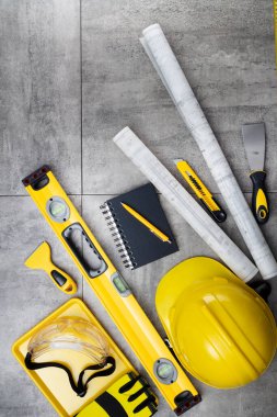 Müteahhit teması. Müteahhidin alet çantası, sarı başlık, libella, el testeresi. Gri karoların arka planındaki planlar ve defterler.