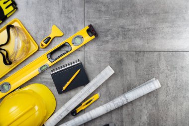 Müteahhit teması. Müteahhidin alet çantası, sarı başlık, libella, el testeresi. Gri karoların arka planındaki planlar ve defterler.