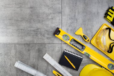 Müteahhit teması. Müteahhidin alet çantası, sarı başlık, libella, el testeresi. Gri karoların arka planındaki planlar ve defterler.