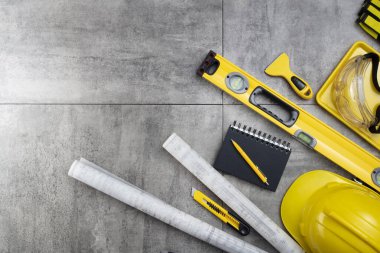 Müteahhit teması. Müteahhidin alet çantası, sarı başlık, libella, el testeresi. Gri karoların arka planındaki planlar ve defterler.