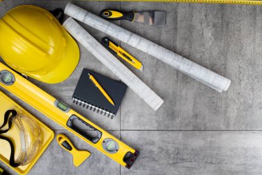 Müteahhit teması. Müteahhidin alet çantası, sarı başlık, libella, el testeresi. Gri karoların arka planındaki planlar ve defterler.