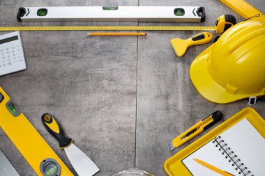 Müteahhit teması. Müteahhidin alet çantası, sarı başlık, libella, el testeresi. Gri karoların arka planındaki planlar ve defterler.