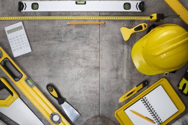 Müteahhit teması. Müteahhidin alet çantası, sarı başlık, libella, el testeresi. Gri karoların arka planındaki planlar ve defterler.