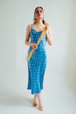 Mavi benekli elbise ve güneş gözlüklü şık bir kız elinde buğday başakları ve stüdyoda poz veriyor.