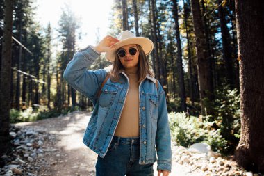 Güz ormanlarında gezen şık şapkalı, kot elbiseli, güneş gözlüklü güzel bir kız.