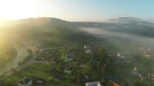 Pueblo, montañas, pequeño río y niebla matutina en los rayos de sol. Disparo aéreo — Vídeos de Stock