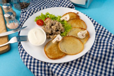 Deruny, mantarlı patatesli gözleme, şampiyonlar, beyaz tabakta ekşi krema. Kirazlı domates, salata. Restoran mavi arka planda servis ediliyor, peçete, çatal, bıçak. Üst görünüm, alanı kopyala