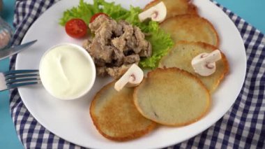 Deruny - mantarlı patates gözlemesi, beyaz tabakta ekşi krema. Kirazlı domates, salata. Restoran mavi arka planda servis ediliyor, peçete, çatal, bıçak. Krep sebze yemeği.