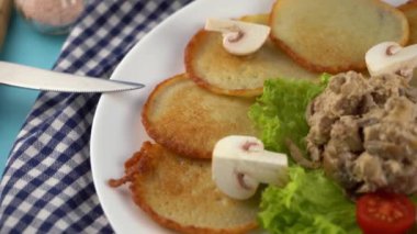 Deruny - mantarlı patates gözlemesi, beyaz tabakta ekşi krema. Kirazlı domates, salata. Restoran mavi arka planda servis ediliyor, peçete, çatal, bıçak. Krep sebze yemeği.
