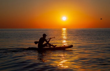 Küreğiyle sörf tahtasında yüzen bir adamın silueti. Denizde şafak güneşi. Bezelye tahtasında yüzmek. Su sporları. Güzel manzara. Denizde gün batımı. Okyanus kıyısı.