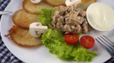 Deruny - mantarlı patates gözlemesi, beyaz tabakta ekşi krema. Kirazlı domates, salata. Restoran mavi arka planda servis ediliyor, peçete, çatal, bıçak. Krep sebze yemeği.