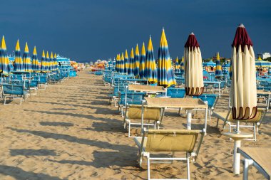 Şemsiyelerle kumlu sahilde güneşleniyorlar. Boş sahil beldesi. İtalya. Sahil şeridi şezlongunda güneş banyosu. Yazın mavi yağmurlu gökyüzü. Rimini.
