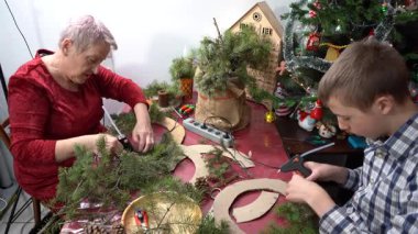 Büyükannem ve torunum birlikte Noel çelengi yapıyorlar. Yapıştırıcı ladin dalları. Yaşlı kadın ve genç çocuk kendi elleriyle dekore ederler. Aile mesleği