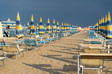 Uzun kumlu sahilde şemsiyelerle güneşleniyorlar. Boş sahil beldesi. İtalya. Sahil şeridi şezlongunda güneş banyosu. Yazın mavi yağmurlu gökyüzü