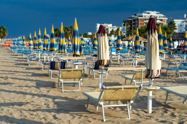 Şemsiyelerle kumlu sahilde güneşleniyorlar. Boş Rimini tatil köyü. İtalya. Sahil şeridi şezlongunda güneş banyosu. Yazın mavi yağmurlu gökyüzü