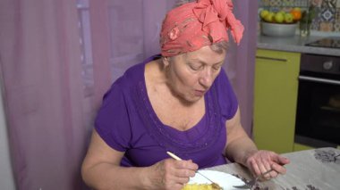 Evde çatal ve bıçakla yemek yiyen yaşlı bir kadının portresi.