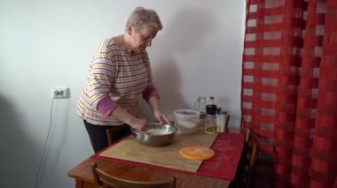 Yaşlı bir kadın hamur yoğurur. Masada un var.
