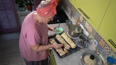 Evde yemek pişiren yaşlı bir kadın. Turtaların tepesini yumurta ile fırçala.