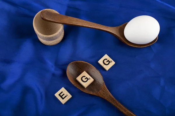 Egyetlen nyers tojás fakanalakkal és fából készült betűkkel kék selyemruhán — Stock Fotó