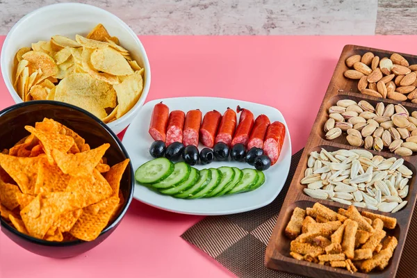 Různé svačinky na růžovém stole. Chipsy, klobásy, pistácie, olivy — Stock fotografie