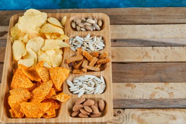 Tahta bir masada çeşitli atıştırmalıklar tabağı. Cips, kraker, badem, fıstık, ayçekirdeği.