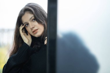 Siyah kazaklı esmer kız pencerenin yanında duruyor.