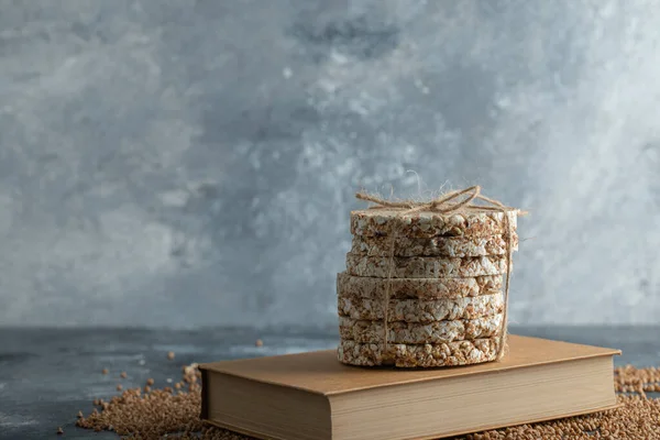 Delicioso pan crujiente, trigo sarraceno sin cocer y libro sobre superficie de mármol —  Fotos de Stock