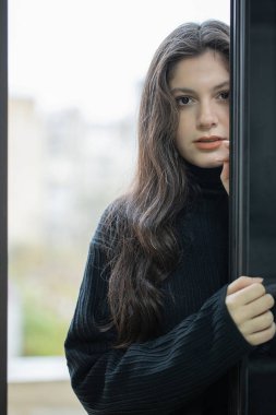 Siyah kazaklı esmer kız pencerenin yanında duruyor.